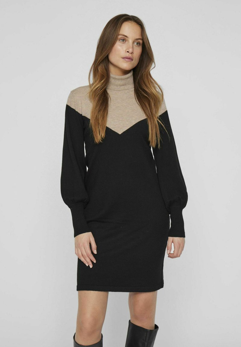 Robe à col roulé noire et beige avec de longues manches bouffantes, présentant un design en bloc de couleurs en chevrons, une texture en tricot douce et une silhouette ajustée.