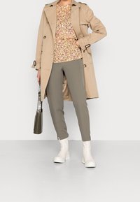 Cappotto beige sopra una maglietta a fantasia floreale, abbinato a pantaloni verde oliva e stivali di gomma bianchi. Include una borsa verde strutturata.