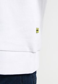Witte sweatshirt met geribbelde zoom, met een klein geel label met een zwart logo aan de zijkant. Het materiaal lijkt zacht en gestructureerd.