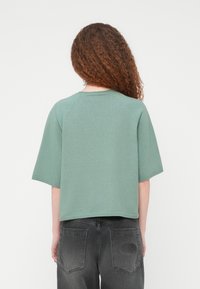 Άτομο με μακριά σγουρά μαλλιά φοράει χαλαρό πράσινο κοντομάνικο μπλουζάκι και ξεβαμμένο μαύρο τζιν, φαίνεται από πίσω σε απλό φόντο.