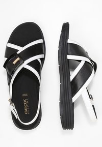 Musta-valkoinen sandaali säädettävällä soljella, ristikkäisillä nauhoilla, kumipohjalla ja Geox-logolla, jossa on sileä nahkapinta.