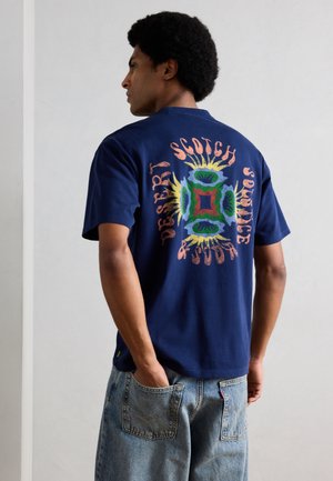 Mężczyzna z afro fryzurą, ubrany w granatowy t-shirt z kolorowym, abstrakcyjnym wzorem i napisem na plecach, w połączeniu z jasnoniebieskimi dżinsami.