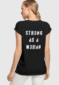 Sort t-shirt med opkørte ærmer og løs pasform. Teksten på ryggen siger "SÅ STÆRK SOM EN KVINDE" i fed, distressed hvid skrift.