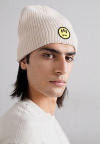 Berretto beige lavorato a maglia con forma arrotondata, caratterizzato da una patch con un sorriso nero e giallo, indossato da una persona con lunghi capelli scuri.