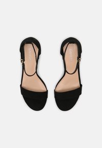 Svarta mocka klacksandaler med rem vid ankeln, bred framdel och en mjuk beige innersula. Har ett gyllene spänne för justering.