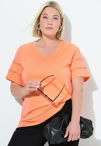 Oranžové tričko s výstřihem do V a texturovanými rukávy, s výřezovými detaily. V kombinaci s černými kalhotami a černou kabelkou. Minimalistické doplňky.