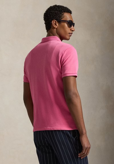 Άνδρας που φοράει μαύρα γυαλιά ηλίου, φωτεινό ροζ κοντομάνικο πόλο πουκάμισο και σκούρο μπλε παντελόνι με λεπτές ρίγες, κοιτάζοντας πλάγια μπροστά σε απλό φόντο.