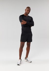 Black long-sleeve athletic shirt, black shorts, and silver running shoes with orange accents. Model poses with arms crossed against a plain background.