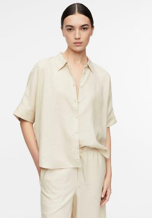 Młoda kobieta o ciemnych włosach, ubrana w beżową krótkomanicową koszulę na guziki i pasujące luźne spodnie, stojąca na białym tle.