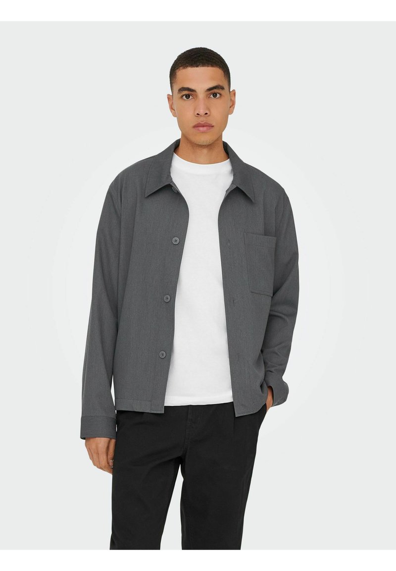 Chemise grise à boutons avec une coupe décontractée, poche avant et texture lisse, associée à un t-shirt blanc à col rond et un pantalon noir.