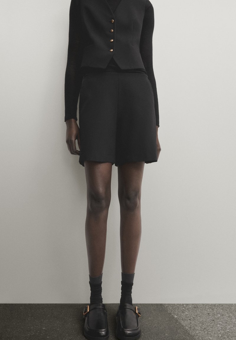 Gilet noir avec des boutons dorés associé à un short noir, accentué par des chaussettes noires et des chaussures slip-on noires dotées d'un détail de boucle marron.