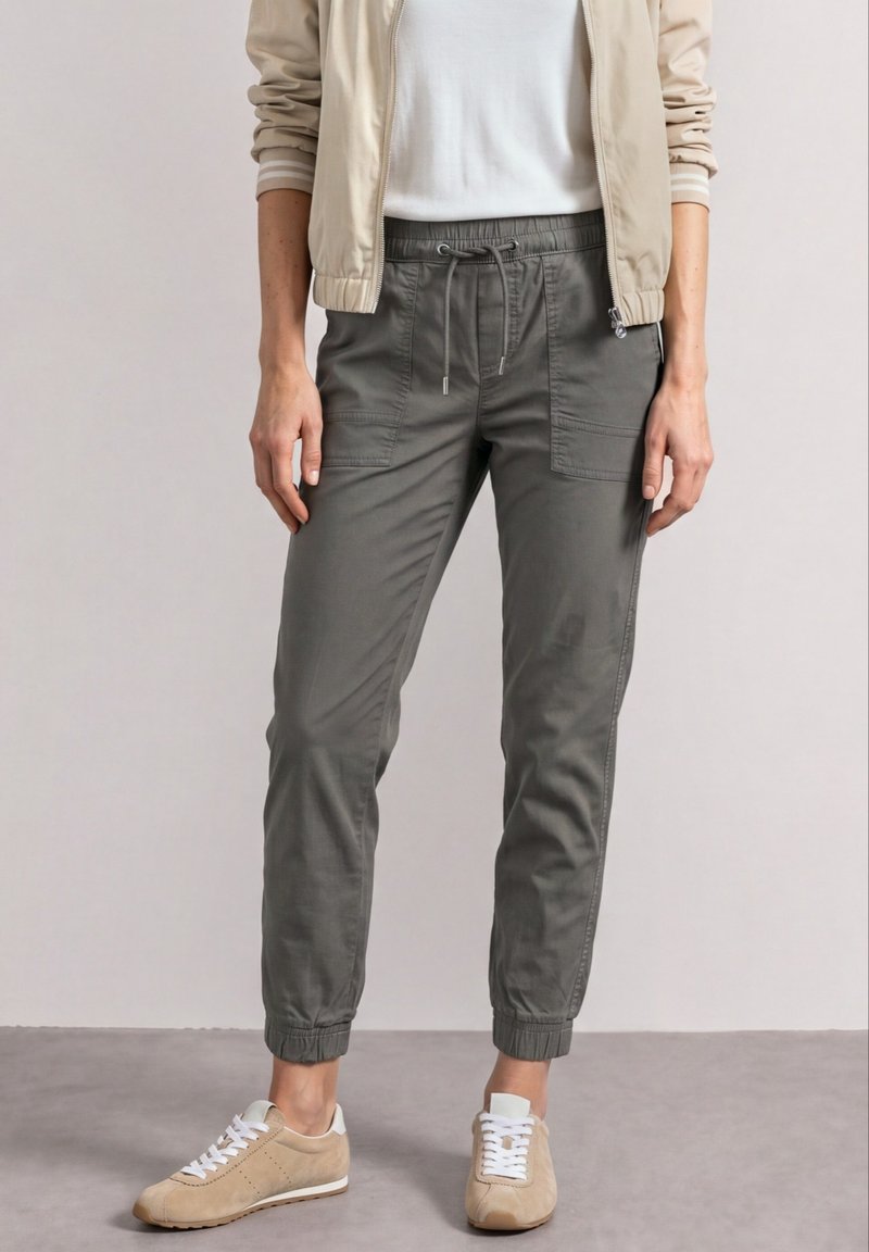 Personne portant un pantalon de jogging gris à cordon, une veste légère beige, un t-shirt blanc et des baskets beiges, debout sur un sol gris.