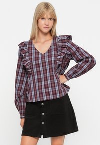 Pruimen- en blauwe geruite blouse met rimpel mouwen, V-hals en lange mouwen; gecombineerd met een zwarte corduroy mini-jurk met knopen aan de voorkant.