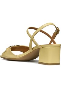 Sandal de cuero amarillo de punta abierta con tacón bloque, que presenta tiras entrecruzadas y un acento decorativo en el área de los dedos. Textura suave.