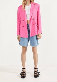 Blazer de color rosa brillante, de doble botonadura, con dos bolsillos frontales, combinado con pantalones cortos de mezclilla con dobladillos deshilachados y sandalias de plataforma negras.