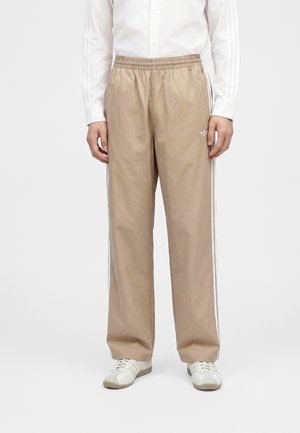 Persona che indossa pantaloni della tuta beige con strisce bianche laterali, sneakers bianche e una maglietta bianca a maniche lunghe, in piedi davanti a uno sfondo semplice.