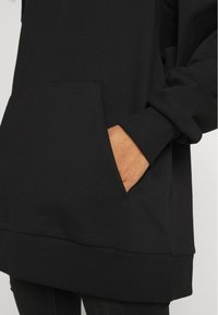 Sudadera negra con un bolsillo tipo canguro, puños y dobladillo de canalé. Tejido suave con una textura lisa, mostrando parte de un brazo con la mano dentro del bolsillo.