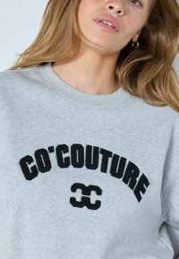 Szürke pulóver kerek nyakkal, vastag, fekete hímzett "CO'COUTURE" felirattal és fekete dupla ívű logóval.