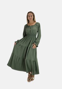 Maxi-kjole med lange ærmer i olivengrøn med smocktalje, lagdelte flæser og knaplukning. Blødt stof og flydende silhuet.
