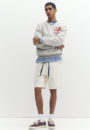 Młody mężczyzna stoi ze skrzyżowanymi ramionami, ubrany w szarą bluzę, niebiesko-białą paskowaną koszulę, białe spodenki, białe skarpetki i brązowe trampki.