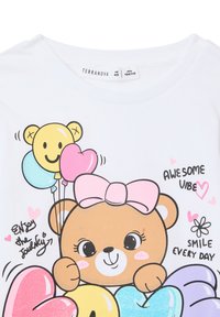 T-shirt bianco con una grafica di un orso che tiene palloncini multicolori, dotato di un fiocco rosa, e elementi testuali come "AWESOME VIBE" e "SMILE EVERY DAY."