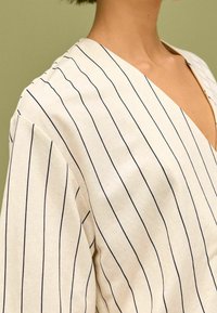 Puuvillainen paita, jossa on V-pääntie ja vaalean valkoisen taustan päällä vaakasuoria tummansinisiä raitoja. Pehmeä rakenne, leveät hihat, näkyvät saumat.