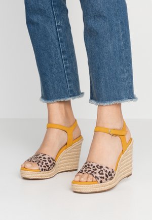 Benen in cropped blauwe jeans met gele wedge sandalen met een luipaardprint op de voorkant en gevlochten touwen zolen op een witte vloer.