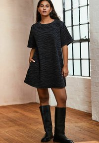 Robe t-shirt en denim noir avec un motif zèbre subtil, manches courtes, encolure ronde et poches latérales ; associée à des bottes noires montant jusqu'au genou.