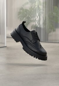 Zwarte leren brogueschoen met dikke grof profielzool, vleugelvormige details en veters, zwevend boven een betonnen vloer met een achtergrond van metalen verticale jaloezieën.