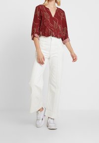 Blouse bordeaux à motif avec un col en V et des manches trois-quarts, associée à un pantalon large taille haute blanc et des baskets grises.