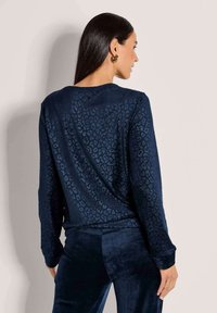 Vrouw met lang donker haar, die gouden oorringen draagt en een langslavend diepblauwe leopardprint bovenstuk met bijpassende broek, gezien van achteren.