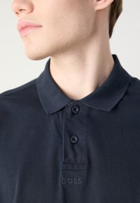 Camisa polo azul-marinho em algodão, com colarinho, três botões e um logótipo bordado "BOSS" no peito. Textura subtil visível.