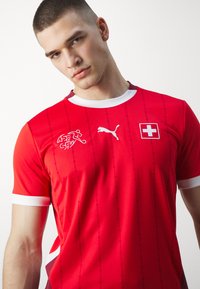 Rød atletisk trøje lavet af åndbart stof. Har hvide accenter på kraven og ærmerne, Schweiz' flag og Puma-logoet på brystet.