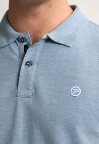 Světle modré polo tričko se strukturovanou tkaninou, dvěma knoflíky a kulatým logem na levém prsu. Klasický límec se jemným prošíváním.