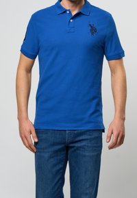 Muž v modrém polo tričku s krátkým rukávem a malým vyšitým logem na hrudi, v modrých džínách, stojí s uvolněnýma rukama.