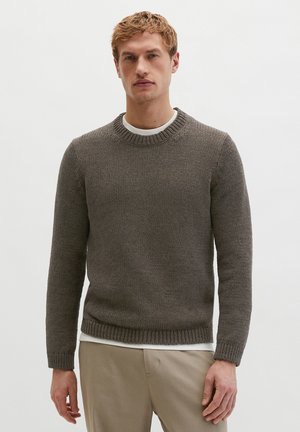 Junger Mann, der einen grauen Strickpullover über einem weißen Hemd und beigen Hosen trägt, vor einem schlichten hellen Hintergrund stehend.