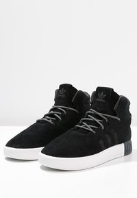 Baskets montantes en velours noir avec une semelle en caoutchouc blanc, présentant des détails texturés et des lacets contrastants. Logo à trois bandes sur le côté.