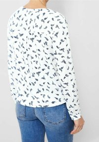 Person iført hvid langærmet bluse med marineblåt paisley mønster og bløde jeans, vist fra ryggen mod en ensfarvet baggrund.