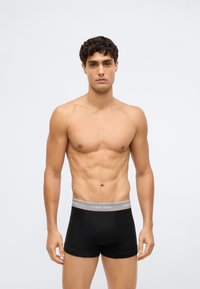 Homem sem camisa, com cabelo curto e encaracolado, vestindo boxers pretos da Calvin Klein, de pé contra um fundo claro e liso.