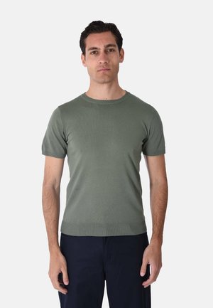 Uomo con capelli corti e scuri che indossa una maglia a maniche corte verde oliva lavorata a maglia e pantaloni scuri, in piedi con espressione neutra su sfondo bianco.