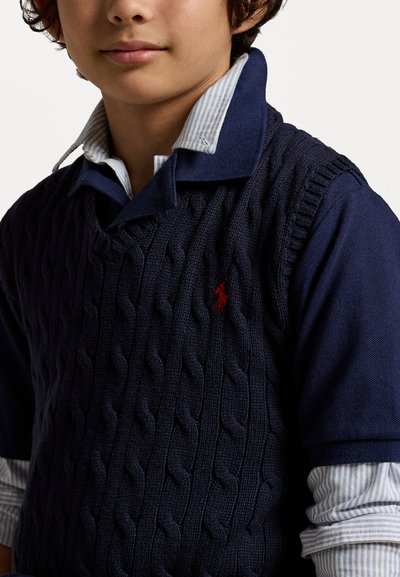 Ναυτική πλεκτή γιλέκ�ο με σχέδιο καλωδίωσης πάνω από ένα σκούρο μπλε πουκάμισο με λεπτές λευκές ρίγες. Κόκκινο λογότυπο στη γιλέκο.