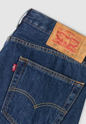 Vaqueros Levi's de mezclilla azul oscuro con bolsillo trasero visible, costuras amarillas, etiqueta roja de Levi's y etiqueta de cuero marrón de la marca con detalles de tallas.