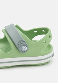 Vihreät muoviset sandaalit, joissa on harmaa hihna, jossa on pyöreä emblem, valkoinen välipohjan raita ja teksturoitu pohja pitoa varten.