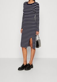 Marinrandig ribbad klänning med rund hals och långa ärmar; har en slits på sidan. Kombinerad med svarta grova loafers och en liten handväska.