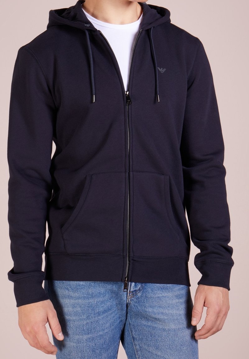 Marinblå zip-up hoodie i ett mjukt tyg, med dragsko i huvan, framfickor och subtil logotypdetalj på bröstet.