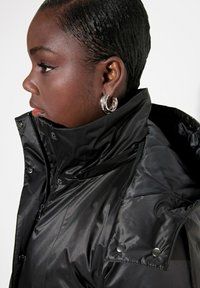 Giubbotto puffer nero con cappuccio, colletto alto, texture lucida, dettagli con bottoni a pressione e chiusura laterale con zip. La modella indossa orecchini a cerchio argentati.