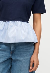 Άτομο που φοράει μπλε σκούρο κοντομάνικο τοπ με λεπτομέρεια πεπλουμ με ανοιχτό μπλε και λευκές ρίγες και μεσαίου τόνου μπλε τζιν.