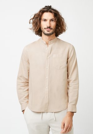 Homme aux cheveux bouclés mi-longs, portant une chemise beige à boutons avec col mandarin et un pantalon blanc, debout sur un fond blanc.