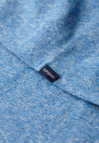 Blauw gemêleerde stof met een genaaide marineblauwe geweven label waarop de merknaam "Superdry" in witte tekst staat.