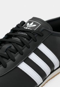 Detailní záběr na černé tenisky Adidas s bílými pruhy, černými tkaničkami a logem na jazyku, ukazující část gumové podrážky.
